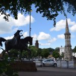 A traffic junction, with a pointed stone market cross at the centre of a roundabout. To the left, in the foreground is a state of a woman on a horse, mostly in silhouette. Trees overhang the picture. Various buildings, including a church tower, are in the background.