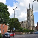 A grey stone church with a grey clock tower, with turrets on its corners. It sits on a s mall graveyard on a wide street, with brick buildings either side. There are trees on the street.