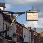 A sign widely overhangs a street, reading “YE OLDE REINE DEER INN. HOOK NORTON ALES”. The sign is supported by a triangle of black beams, on the diagonal one of which are written in gold “HOOK NORTON ALES”. The street below is of red-brick buildings, some of which are painted white, only visible from first floor level upwards. The sky with blue, with white and grey clouds.
