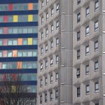 Two tower blocks, photographer so neither top nor bottom is visible of either. The one in front is beige and drab, made mostly of concrete; the one behind has blue strips between floors, and bright red, orange and yellow panels replacing some windows.