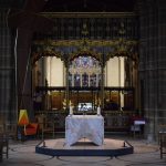A section of a cathedral, with a large tomb in the centre of light stone on a dark plinth. Behind it is a reredos, and another, more brightly-lit chapel is visible behind that.