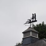 A weathervane, showing to people and various birds silhouetted against a grey sky, on the top of a small turret in front of trees.