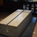 A large block of light stone, trapezoidal in shape, with a deep cross cut into it, sits on a dark plinth on a red-tiled floor, forming a tomb. It is spotlit; behind it, darkened, is a low gate.