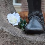 A metal foot of a statue, standing on stone; the foot is shoed, with a visible buckle. Next to it, resting against the stone, is a white rose.