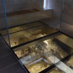 A modern building containing a glass floor, with an archaeological trench beneath it. In one portion the glass floor is removed, and this section is surrounded by four floor-to-ceiling glass walls.