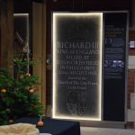 A stone reading “RICHARD III”, “KING OF ENGLAND”, “KILLED AT BOSWORTH FIELD IN THIS COUNTY”, “22ND AUGUST 1485”, “Buried in the Church of the Grey Friars in this Parish”. The stone is about six feet tall and two or three wide; above the text inscription is engraved a crown, and below the Plantagenet coat of arms. The stone is lit around the sides, and there is an information panel (“titled “KING RICHARD III MEMORIAL STONE”) next to it. The stone is in a room containing a Christmas tree which has fallen over.