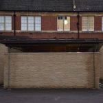 A low building extends out from a wall, both made of light stone; the building has a row of windows around the top. Behind is visible the top row of windows of a brick building with a slate roof.