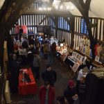 A view down from a balcony into an old timbered wall, with small leaded windows are a high level. There are craft stalls on the ground floor, with people milling around.