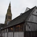 A half-timbered building, with one storey but a high roof, behind which rises a high church tower with a spire. A street leads off to the left.