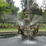 A fountain with three figures, set in a round pond. Behind are grass, shrubbery and trees.