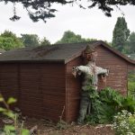A scarecrow against a wooden shed, surrounded by scrubby allotment-like plants. There is a tree overhanging from the left.