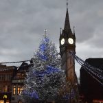 A freestanding clock tower, with a brightly light clock, next to which is a white Christmas tree with white lights. This sits in a city square, under a foreboding sky.