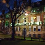 The Town hall in Leicester, bedecked with Christmas lights (including, in green blackletter font, the words “Christmas Greetings”). In front is a square with bare trees.