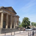 A one-storey building with a portico, the portico having plain Doric columns and steps leading up to it. The building is of Cotswold stone, Behind is a tree-lined market square, with parked cars.