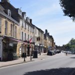 A row of shops along a market square, low buildings, many of Cotswold stone. Many cars are parked in the market square, but the pavements are quiet.