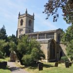 A church, with a square castellated tower and a near-continuous clerestory. The church is of Cotswold stone, and sits in a graveyard with many small trees and a path running through it.