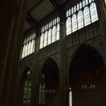 A dimly-lit church nave, with light streaming in through near-continuous clerestory windows. The ceiling has wooden beams. Below the clerestory are gothic arches opening onto the aisle, with a stained-glass window visible at the east end of the aisle.