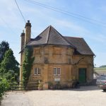 A small two-storey building, in plan like an octagon with a rectangle attached. The octagonal section has a roof rising to a point, with the upper storey having no windows, and instead various reliefs, including a square and compass. The lower storey has small windows. The building is of Cotswold stone, and sits in front of trees and rolling hills. There is a car parked in the building’s bare yard.