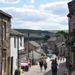 Looking down a steep, slightly-winding cobbled street, with yellow-stone buildings (the odd one painted white, most soot-blackened) on either side. There are many people walking up the street, and several cars parked alongside. The buildings are mostly shops, though there are some private houses, one in the foreground with bright hanging baskets. There are steep hills in the background, one of which is topped with wind turbines.
