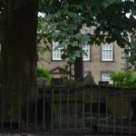 A view towards a two-storey soot-blackened stone building with large sash windows, seen though a dark churchyard with thick tree cover.