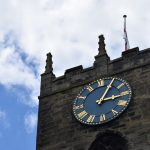 A church tower, of dark stone, with castellation and small spires at the top. There is a dark blue clock with gold hands and Roman numerals on the tower wall. There is a flag pole on top of the church with a furled England flag. A partly-cloudy sky is behind the tower, with some tree leaves poking into frame from the left.