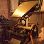 A Linotype machine in a gallery, which has a brick wall and a large shutter covering a window hole.