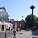 A street view, mostly empty, with one person walking across the foreground. On the left of the street is a metal-framed building with a hipped roof, beneath which is a large window. On the right is a brick warehouse, and a large metal pole with a cog wheel on top. Brick warehouses are at the end of the street.