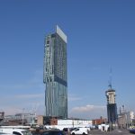 A tall glass skyscraper, which is rectangular in plan, and has a cantilevered overhang about half way up (making the floors a larger rectangle). On the opposite site from the overhang the building has a large grille placed like a billboard above its top storey. The surrounding buildings are mostly low-rise, and there is a car park in the foreground. The sky is mostly blue, with some clouds near the horizon.