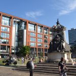 A statue of Queen Victoria, seated, on a plinth with steps. To the left is a brick building with large windows, modern, and a 1960s office block is to the right; these form part of two sides of a public square, grassed, with many people in it.