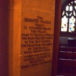 Engraved into a stone pillar in a church is black writing reading “THE BRONTË FAMILY VAULT IS SITUATED BELOW THIS PILLAR, NEAR TO THE PLACE WHERE THE BRONTËS’ PEW STOOF IN THE OLD CHURCH. THE FOLLOWING MEMBERS OF THE FAMILY WERE BURIED HERE MARIA AND PATRICK. MARIA, ELIZABETH, BRANWELL, EMILY JANE, CHARLOTTE. Behind the pillar are visible three rows of pews, a small altar, and a Gothic stained-glass window.
