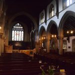 Looking up the nave of a church. There is a row of Gothic arches separating the nave from each aisle. There are small chandeliers illuminating the church, as well as light from the clerestory windows. There is a gothic stained glass window at the east end of the church. The timber sloping ceiling above the aisles is also visible. The walls above the clerestory and along the aisles are painted white, but otherwise the walls are bare stone.