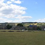 A field, with a dry stone wall across it. There are a few sheep in the field. The grass is short and dry. At the far side is a road, with small semi-detached houses of soot-blackened stone along it. Behind are yellow fields on a hill, with patches of sunlight.