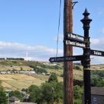A black metal signpost with four arms, reading “Bronte Parsonage”, “Tourist Information” and “Railway Station” on the arms pointing to the left, and “Toilets” on the one to the right. The land falls away behind the signpost into a valley, and on the other side are fields on a hill, which is topped by a wind turbine. There are a few houses in the foreground and a few on the hill. Just behind the signpost is a wooden telegraph pole. The sky is blue, with a few white clouds.