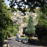 Looking down road on a steep hill. There are trees along the road and overhanging the game, and cars parked along the road, with houses alongside, built of stone. On the far side of the valley is a hill, with buildings (mostly houses) on the hillside to near the top.