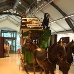 An antique horse-drawn tram, on rails, set into the floor of a museum. There are various figures on the tram, and a sign on its front reading “GREENWICH & WATERLOO STN”. The museum floor is quiet, although there are a couple of people near the lift in the background.