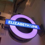 A roundel with a purple circle and a blue bar, reading “ELIZABETH LINE”. It is suspended from a wall. Behind it is the title of an exhibition “SECRET LIFE OF A [obscured]GAPROJECT with some images dotted onto a white wall.