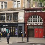 Looking across a street, with a Pedestrian crossing, to a shop which is next to a disused Underground station. The station fascia is of oxblood-coloured tile, with a large arched window above the boarded-up door. Above the arched window is text reading “PICCADILLY RLY” and below is “STRAND STATION”, in white on black. The shop next door says “Language Centre” above the window. The pavement is busy.