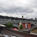A building spans the length of a bridge across a river. The bridge has stone piers and blue-and-white metal arches. The building has glass walls and roof fins. Next to every bridge pier are two columns in the water, painted red. In front of the river is a road which passes under the bridge, and between these a footpath on which are a group of workers in high-visibility clothing.