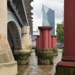 Looking along the side of a bridge over a murky river. The bridge piers are of yellow stone, between greyish metal arches. A building with a spiky roof crosses the bridge. To the right of each bridge pier is a red column in the water, including one only partially visible in the foreground. There is a second row of these to the right, some of which are visible. A glass skyscraper and a tree can be seen on the far bank.