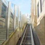 Looking up the track of an inclined lift. To the right is a marble-effect stone building; to the left are glass panels with steps behind. At the head of the track is a glass door, above which the dome of St Paul’s is visible.