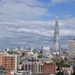 An elevated view of London, dominated by the Shard about two-thirds of the way to the right. Canary Wharf is in the background. The sky has many white fluffy clouds, but is bluer towards the top of the frame.