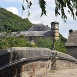 A steeply humped bridge with a cobbled pavement, seen from low down. Behind it is the upper storey of a stone building with a stone roof, with a white window from visible. In the background is the top of a large building with a clock tower, and behind it a steep wooded hill.
