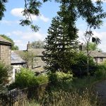 The edge of a churchyard, with a railing along the edge. The yard has overgrown plants and a curving cobbled path. The lane behind the railing, at a slightly lower level, has squat, soot-blackened stone buildings along it.