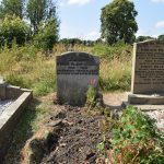 A headstone in a graveyard, reading “IN MEMORY SYLVIA PATH HUGHES 1932 – 1962 EVEN AMIDST FIERCES FLAMES THE GOLDEN LOTUS CAN BE PLANTED”. The word “HUGHES” is partially scratched off. The plot in front is of freshly-tilled soil, with some plant growth and a small red flower. To the left is a grave with no visible writing; a stone for Horace and Emily Draper is on the right. Behind the grayeyard is overgrown and semi-wild. In the background are some trees.