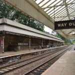 A station, viewed from one of its platforms. The signage is old-style, with a visible sign in the foreground reading “WAY OUT” in white raised text on black, with a hand silhouette pointing to the exit. The canopies on both platforms are old, and the line stretches off into the distance. The platforms are fairly busy.