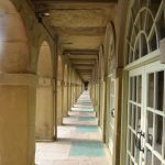 Looking along a stone colonnade. The left-hand side has round arches while the right has matching arched doors and windows. The flagstones on the ground are of worn stone.