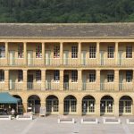 A view from a distance of a three-tiered building, of stone, with a stone roof. The bottom storey has arches leading onto a courtyard, with glazed doors behind. The next has square columns, with simple detailing and railings (pale green) in between, with pale green doors and normal-sized windows on the ball behind. The next gfloor has round Doric columns, with railings, doors and windows as on the floor below. There is a marquee in the courtyard. behind the building a wooded hillside can be seen, with a St George’s flag on the left.