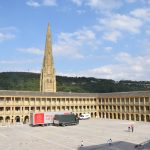 The Piece Hall Yard, seen from a corner. Two sides of a square building surrounding a courtyard are visible, both of three storeys, with colonnades on every tier. The building is of yellow stone, with a grey stone courtyard. A lorry belonging to the BBC’s Antiques Roadshow is in the courtyard. Behind the building is a stone church tower with a very high spire, and in the background a wooded hill beneath a blue sky dotted with clouds.