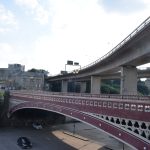A metal bridge, with ornate detailing, painted dark red and cream, flies from the foreground on the right to the background on the left. Above it is a concrete flyover, which flies from above the older bridge’s nearer pier to the background at the centre. Both bridges span a valley containing a car park, with no visible water course. The far side has various buildings, mostly stone, including a tall mill visible under the legs of the flyover.