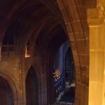 A view from high in the nave of a red-stone cathedral, from the right-hand side balcony. Gothic arches arch over the roof and aisles. There is an internal bridge at the bottom, and some stalls are visible beyond it.